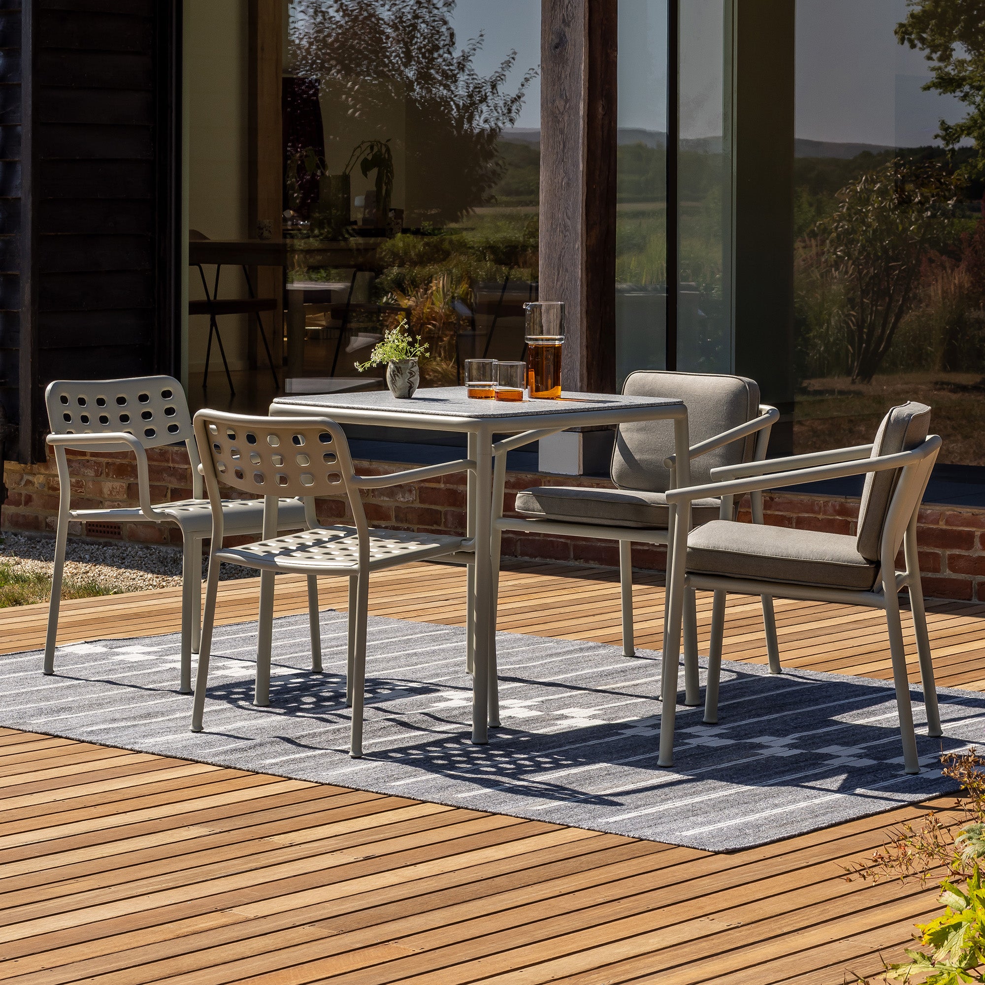 two outdoor Aeros Armchairs and two Campus Armchairs set around a Campus square table in pebble grey with a light granite top. orange glasses and carafe on the table in front of a large glass window.