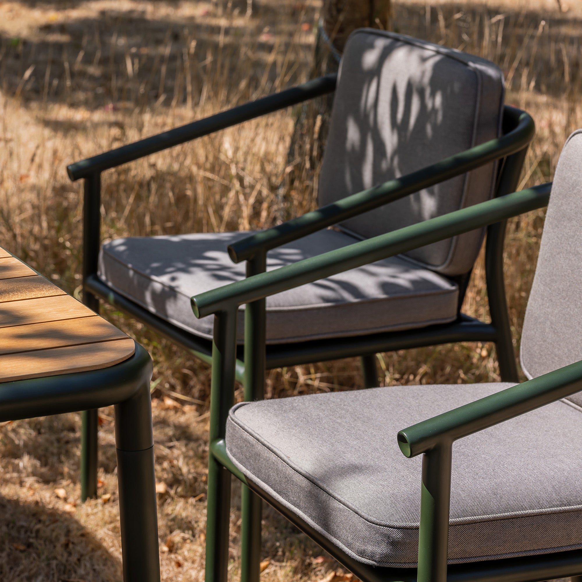 Campus outdoor Dining chair in bottle green powder coat with grey cushions and a matching teak slatted table on a dry grass lawn.