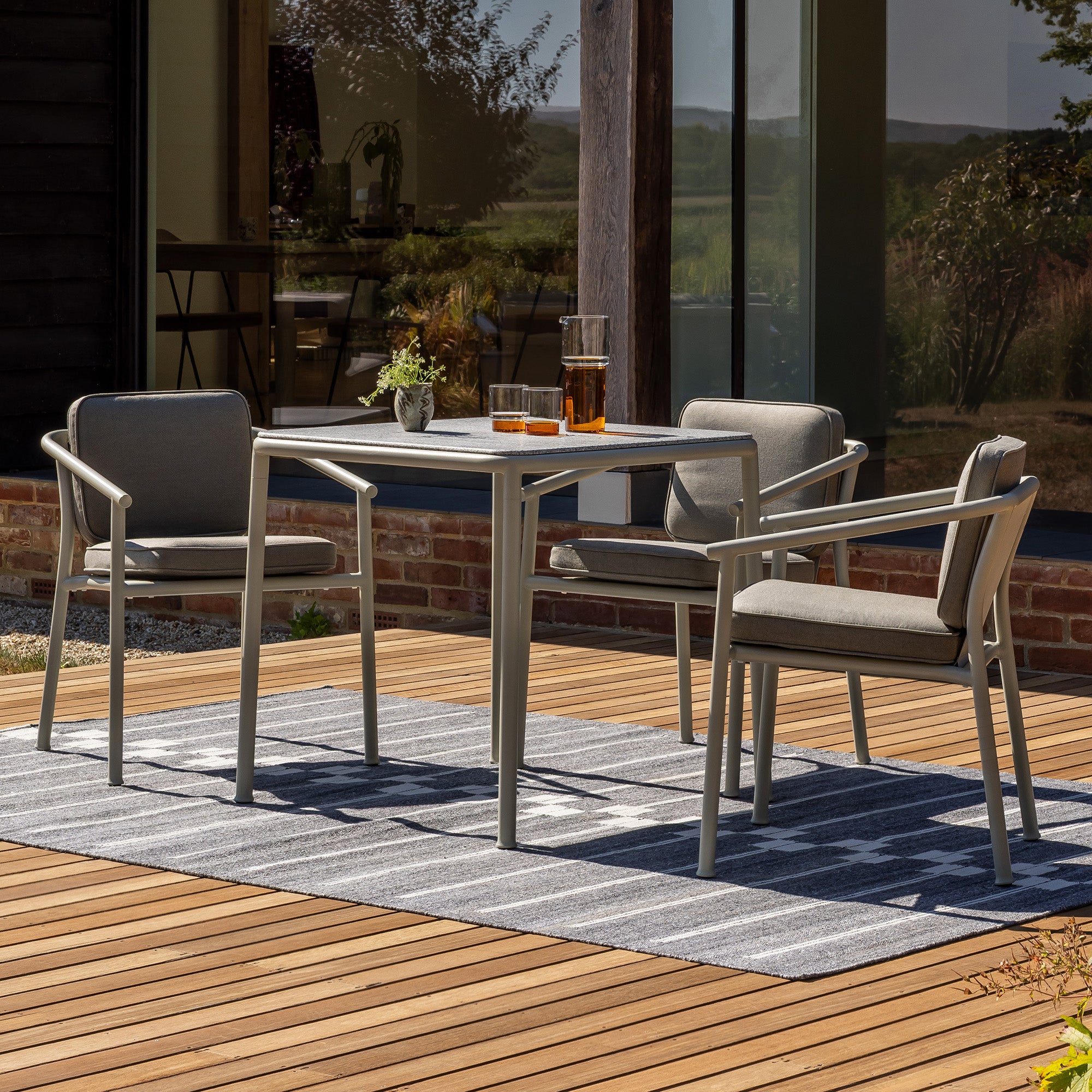 Three Campus armchairs with a Campus square table with granite top on a teak slatted deck with a grey outdoor Spine rug laid below the table in front of a glass window