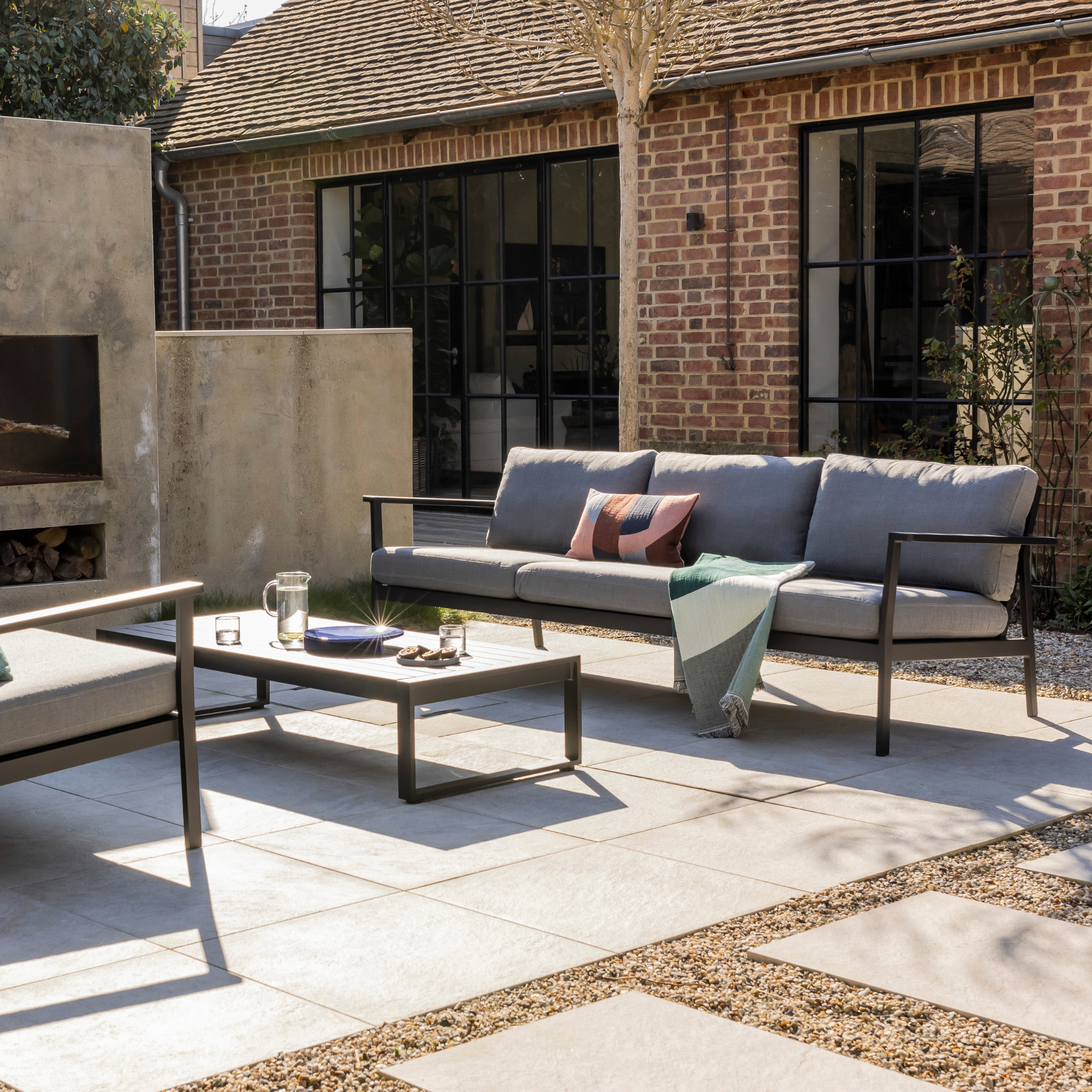 Eos 3 Seat Sofa in Black powder coat with Sunbrella Slate cushions and Eos coffee table in black in front of a concrete outdoor fireplace in a converted barn with crittall windows.