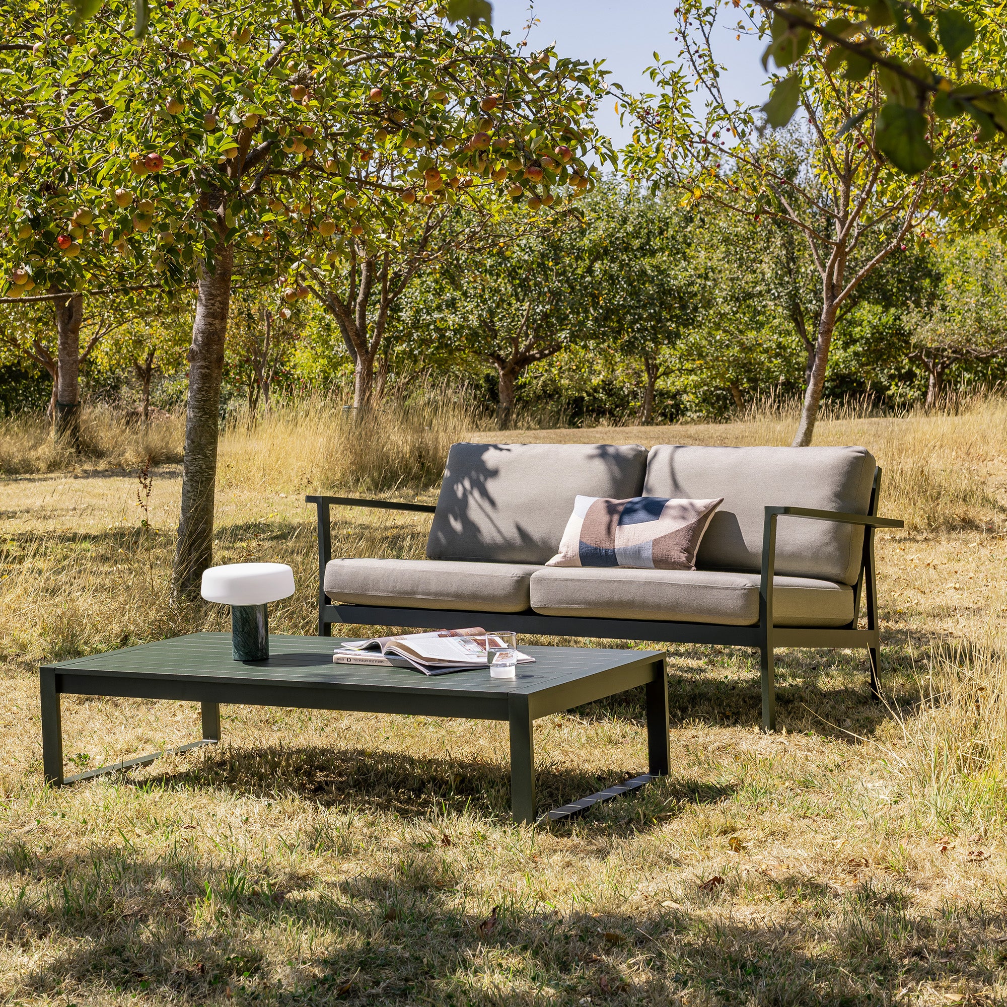 Eos Outdoor 2 seat sofa and coffee table in an orchard under trees, featuring light beige cushions and a dark green powder coat.