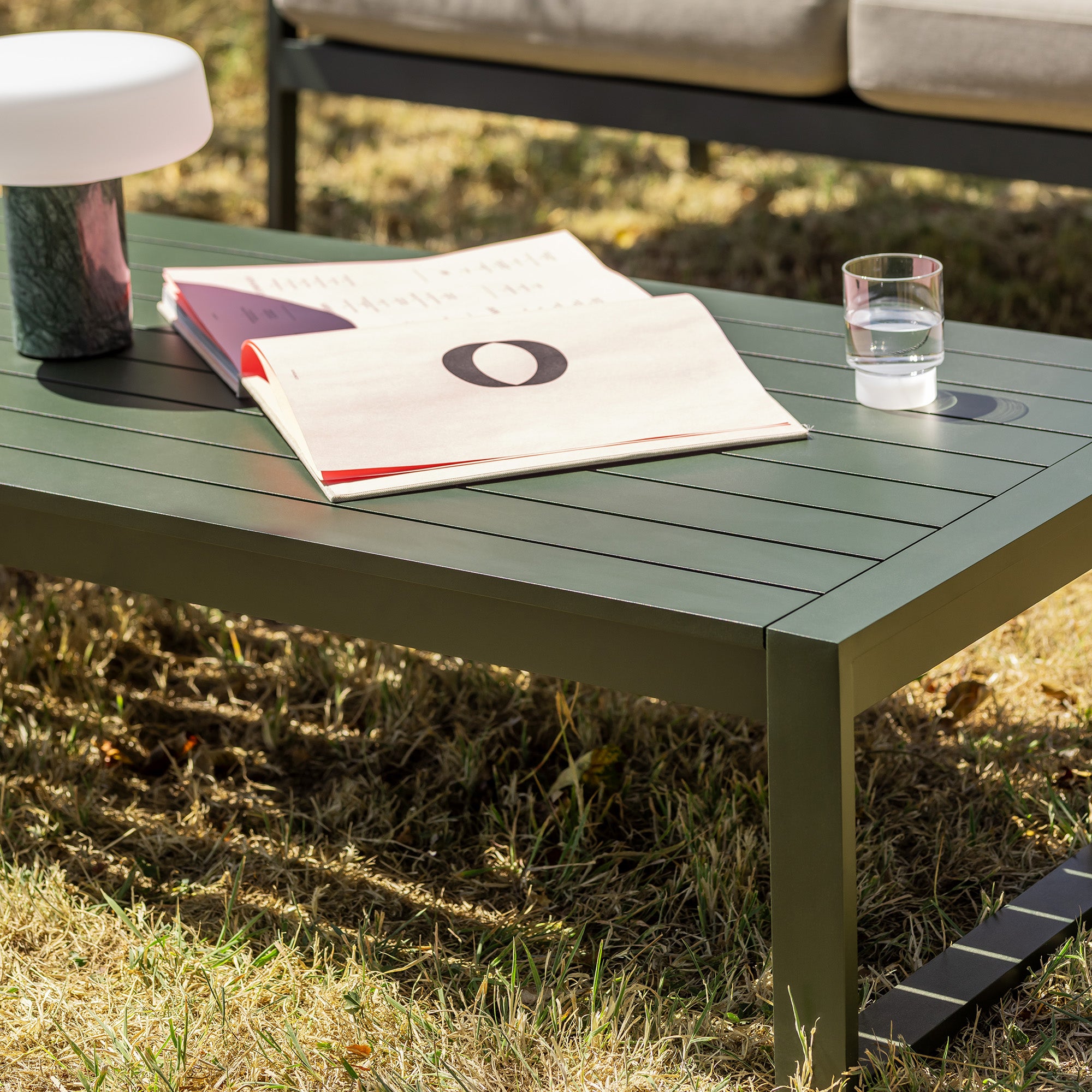 An Eos outdoor coffee table in bottle green, styled with a magazine, a small glass of water, and a modern lamp on a grassy lawn.