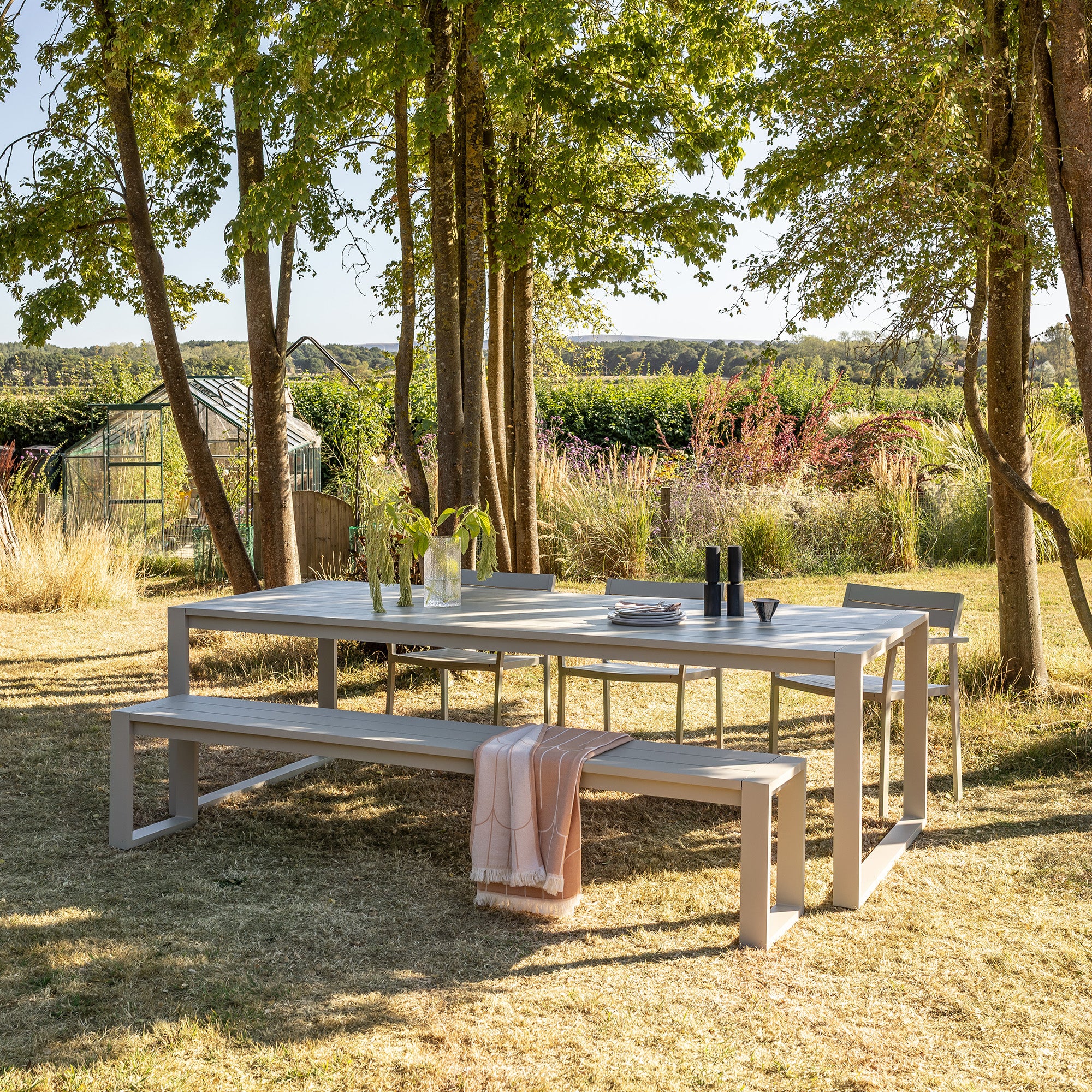An Eos Communal table in pebble grey with a communal bench and three Eos armchairs, arranged on a grassy lawn surrounded by trees in a garden setting.