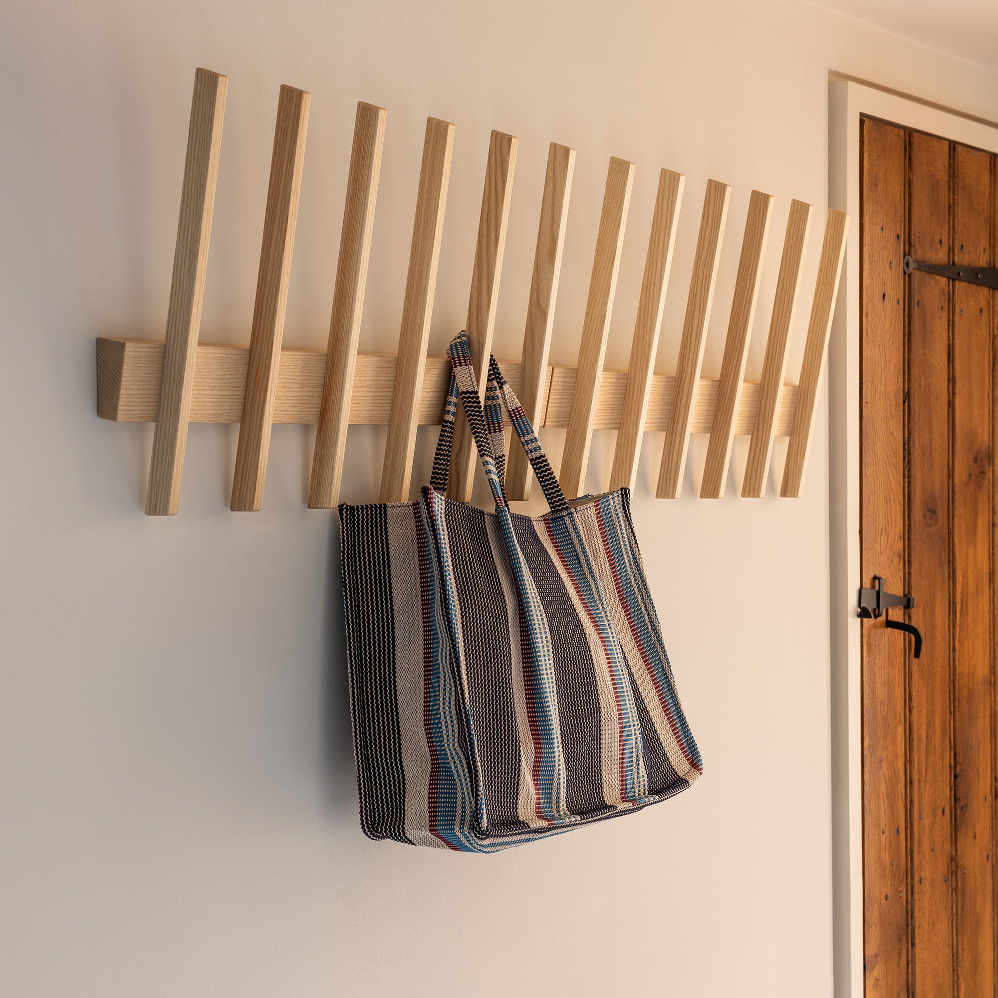 An ash Finn wall coat hook rack mounted on a white wall, with a striped fabric tote bag hanging from one of the hooks next to a wooden door.
