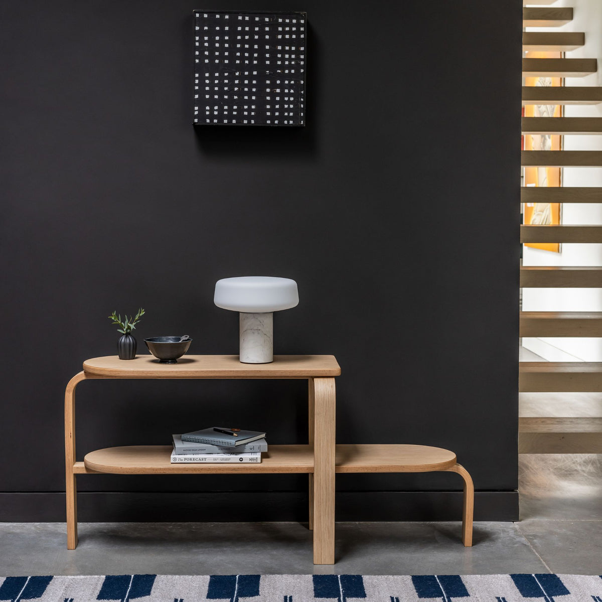 Altura Bench in Oak in a hallway that is painted black with a solid rechargeable light, some books and plant on