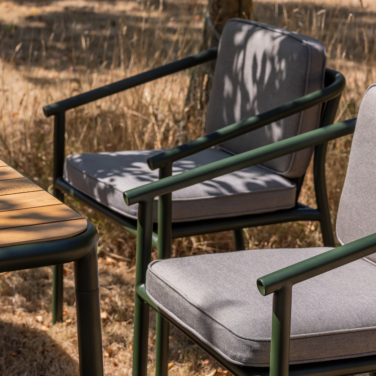 Campus outdoor Dining chair in bottle green powder coat with grey cushions and a matching teak slatted table on a dry grass lawn.