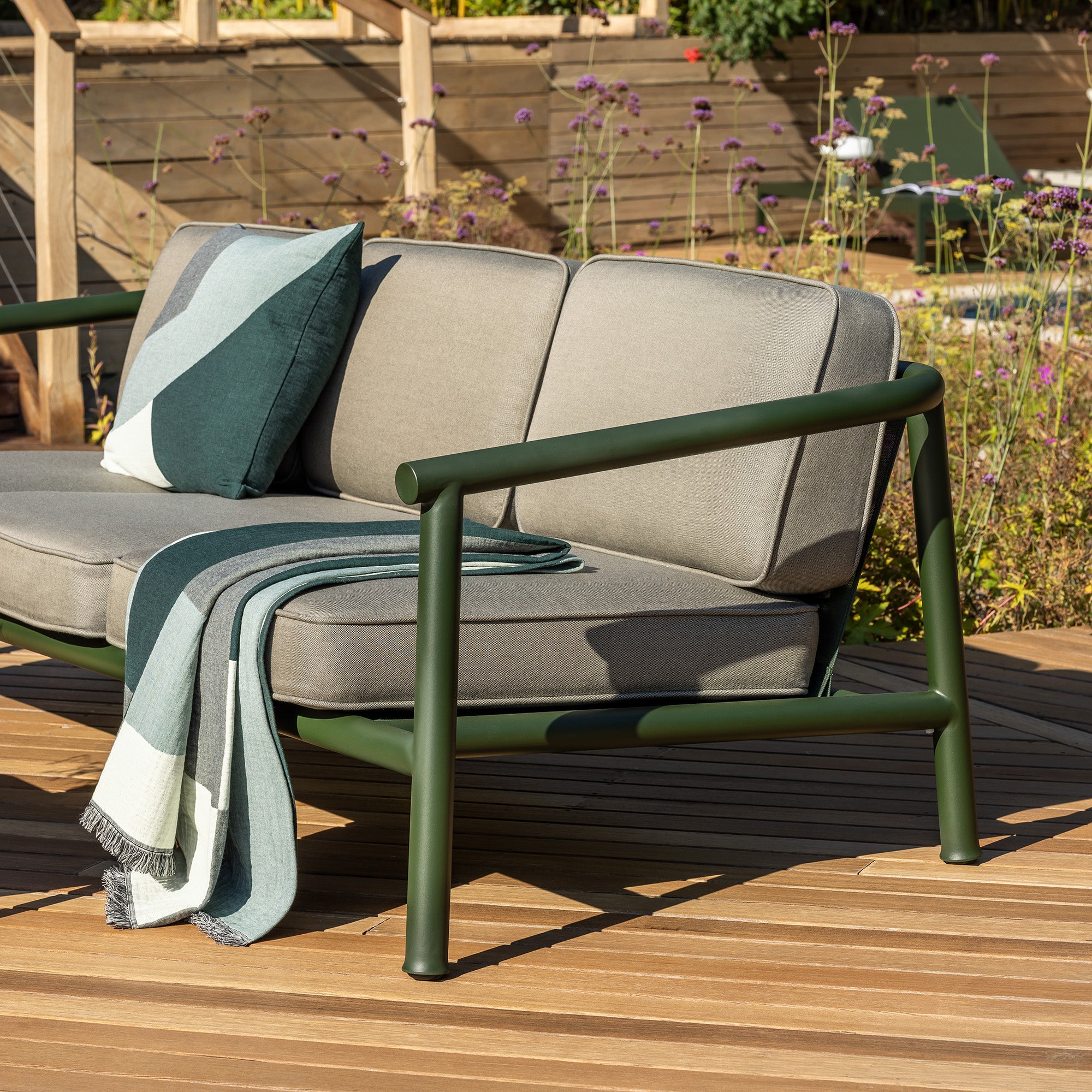 Outdoor Campus two-seat sofa with light grey cushions and a dark green powder coated steel frame, styled on a wooden deck with a matching throw blanket and pillow in a garden setting.