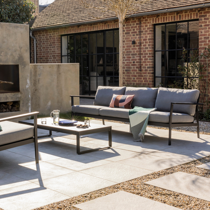 Eos 3 Seat Sofa in Black powder coat with Sunbrella Slate cushions and Eos coffee table in black in front of a concrete outdoor fireplace in a converted barn with crittall windows. 