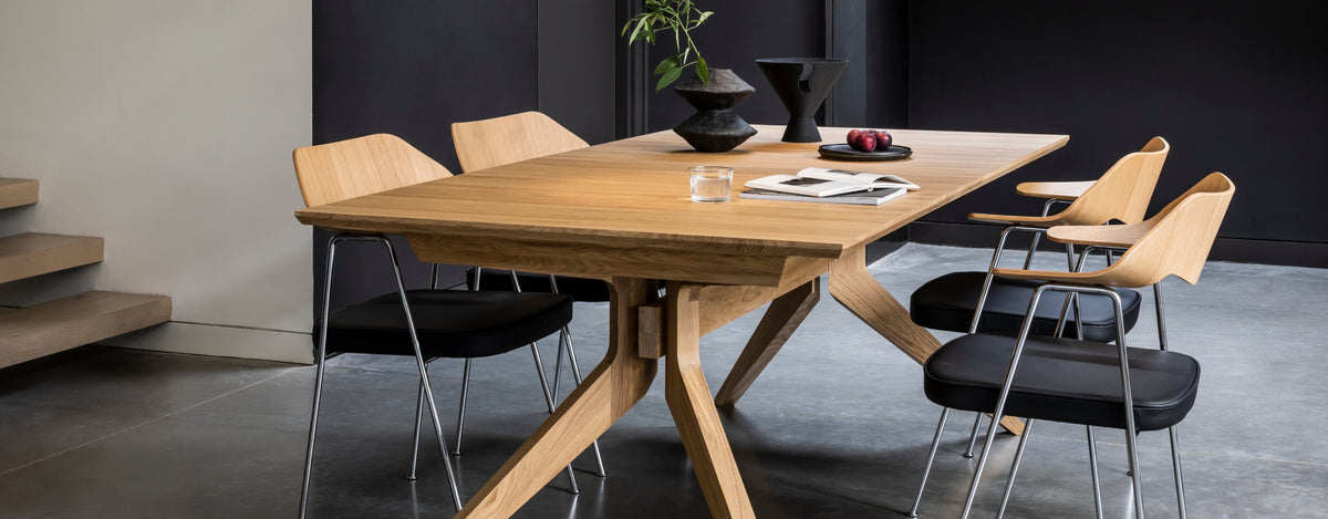 A modern wooden dining table with angled metal legs, surrounded by four chairs with light wood frames and black seats, set in a contemporary dining room.