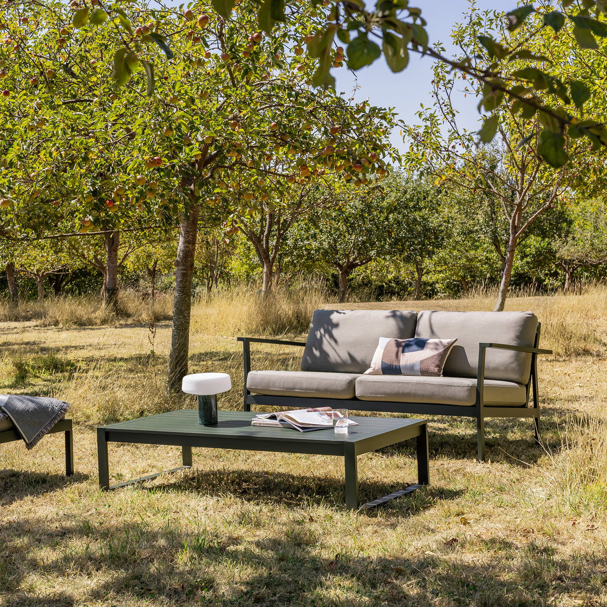 Eos Outdoor 2 seat sofa and coffee table in an orchard under trees with magazines on and a solid rechargeable lamp in Guatemala marble. Sofa has light beige cushions and a dark green powder coat.