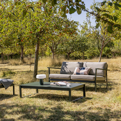 Eos Outdoor 2 seat sofa and coffee table in an orchard under trees with magazines on and a solid rechargeable lamp in Guatemala marble. Sofa has light beige cushions and a dark green powder coat.
