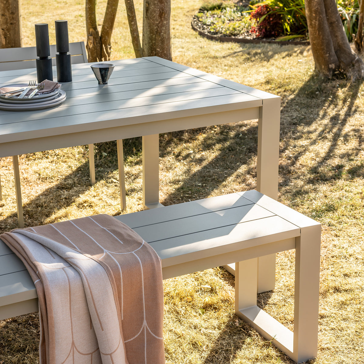 A pebble grey Eos outdoor communal table with communal bench and side chair set on a grassy lawn, styled with plates, cutlery, and a folded peach coloured scales throw.