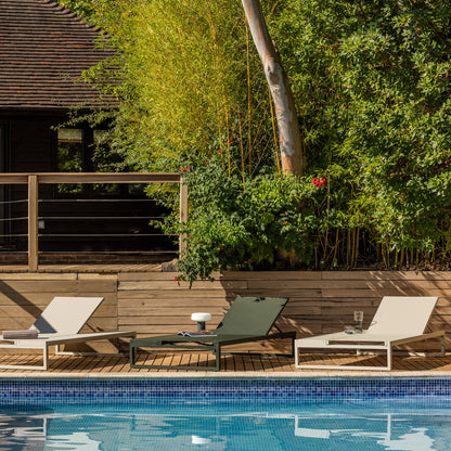 Three Eos outdoor Platform loungers in pebble grey and bottle green arranged on a wooden deck beside a swimming pool, with lush green bamboo and trees in the background. 
