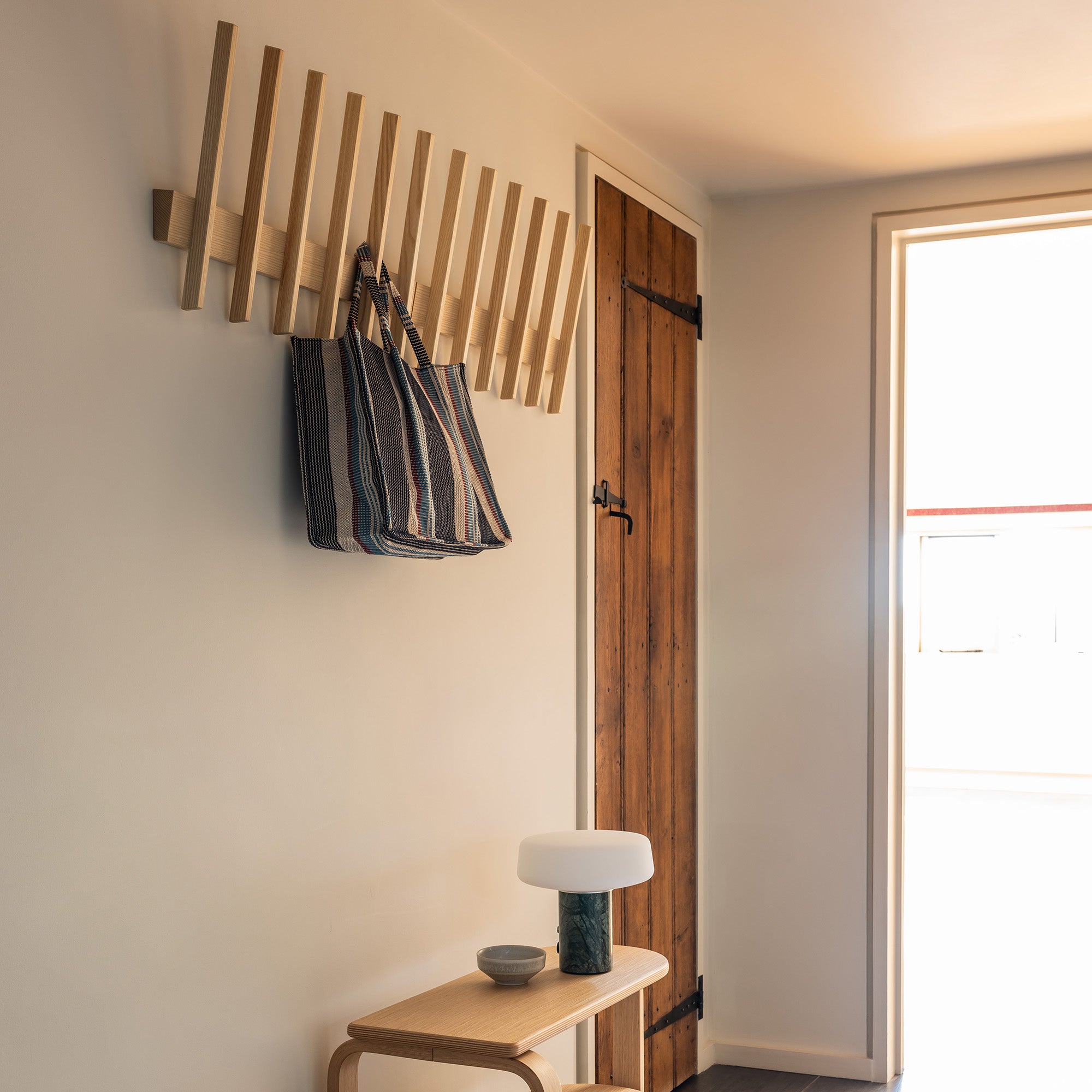 A Finn coat hook in Ash with a striped tote bag on with an Altura bench in Oak beneath in a hallway with a wooden door behind. 