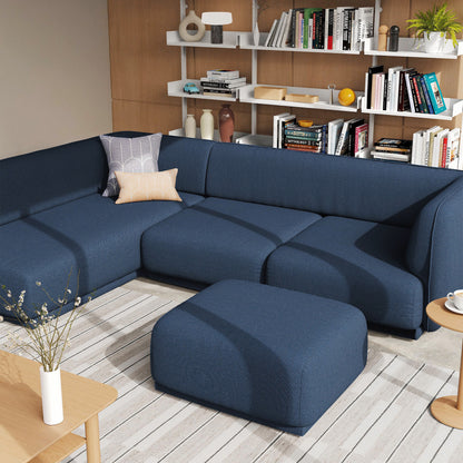 A modern meris sectional sofa in dark blue fabric with a matching ottoman, styled in a contemporary living room setting with a bookshelf and light wood flooring.