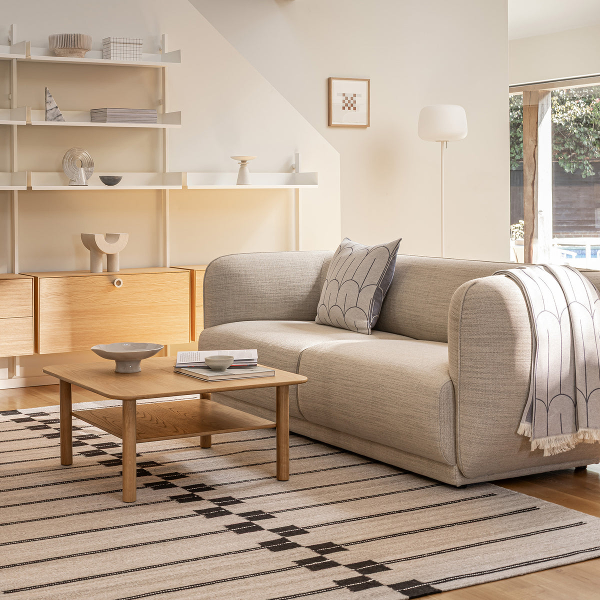 A modern living room scene featuring a light beige upholstered Meris sofa with a scales throw pillow and a throw blanket in blue, set on a striped beige and black Spine rug. An oak Port coffee table sits in front of the sofa, with a white slot shelving with oak cabinets in the background. 