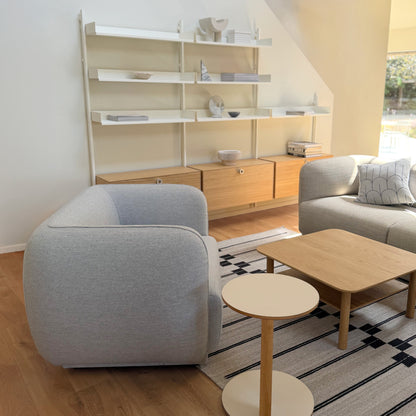 A modern living room setup featuring a light blue Meris armchair in the foreground, a meris 2 seat sofa on the right, an Oak Port square coffee table, a spine rug, and a large white slot shelving unit with cabinets against a white wall.