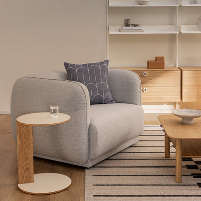 A modern light grey upholstered Meris Armchair designed by Monica Armani  with a blue scales pattern loose pillow, styled in a minimalist living room next to a wooden side table and a striped rug.