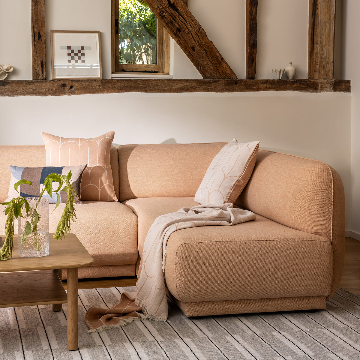 Meris sectional sofa in Blush coloured fabric in a rustic living room setting, styled with peach coloured throw pillows, a cream and peach coloured scales throw, a wooden coffee table, and a warm grey spindle rug.