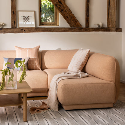 Meris sectional sofa in Blush coloured fabric in a rustic living room setting, styled with peach coloured throw pillows, a cream and peach coloured scales throw, a wooden coffee table, and a warm grey spindle rug.