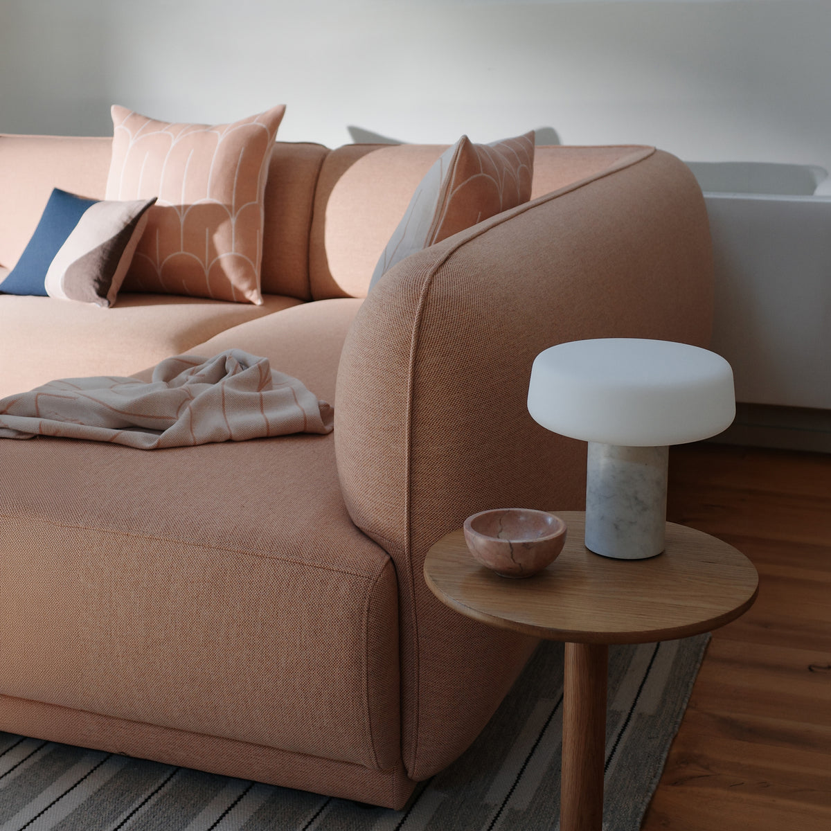 A detail shot of the end of the chaise on the Meris sectional sofa in blush pink fabric in a styled living room setting, featuring scales and eclipse accent pillows, a folded peach scales throw, an Oak Port side table with a white marble solid rechargable table lamp and a marble bowl, and a gray wood floor. 