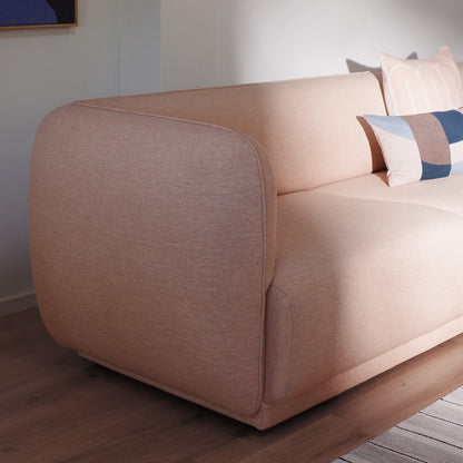 A close-up of the Meris sofa with rounded block arms in a blush fabric, set on a dark wood floor against a light wall with decorative scales and eclipse pillows.