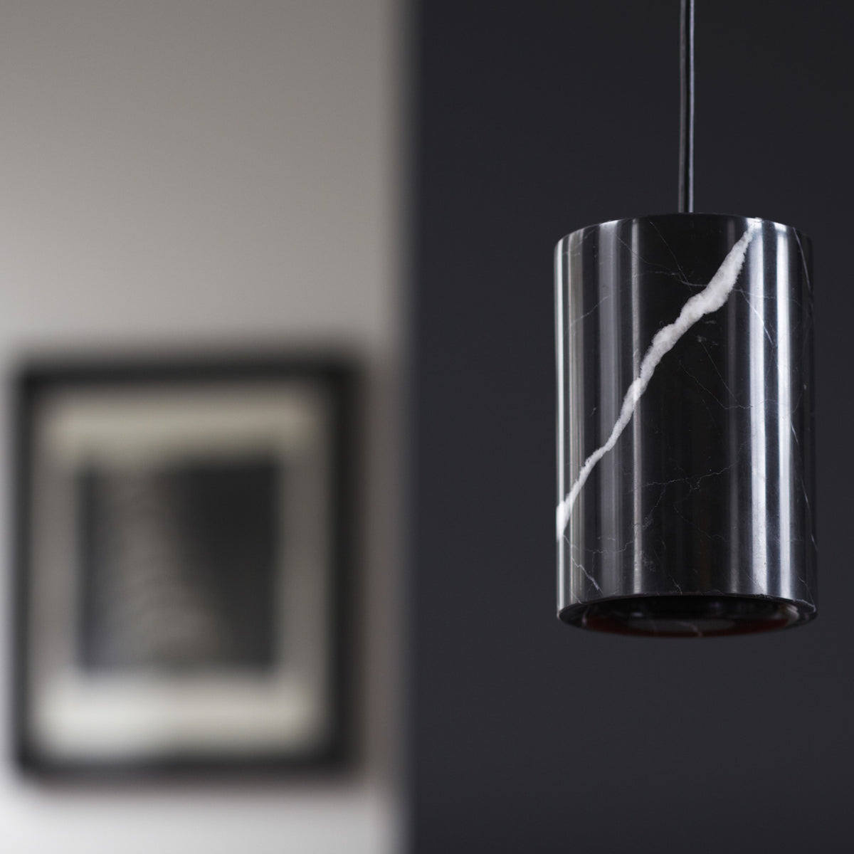 A modern cylindrical pendant light with a glossy black marble finish and a prominent white veining pattern, shown hanging against a dark gray wall.