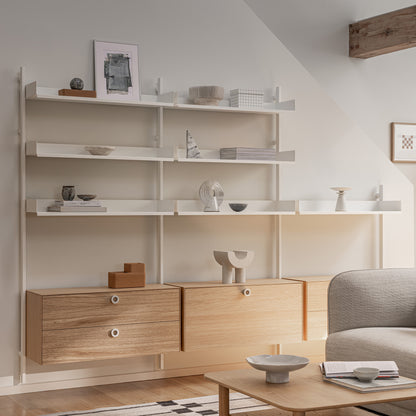Slot shelving in White with both the cabinet and the drawers in Oak mounted on a white wall with minimal books and sculptural items with the corner of a sofa and oak Port coffee table visible in the foreground. 