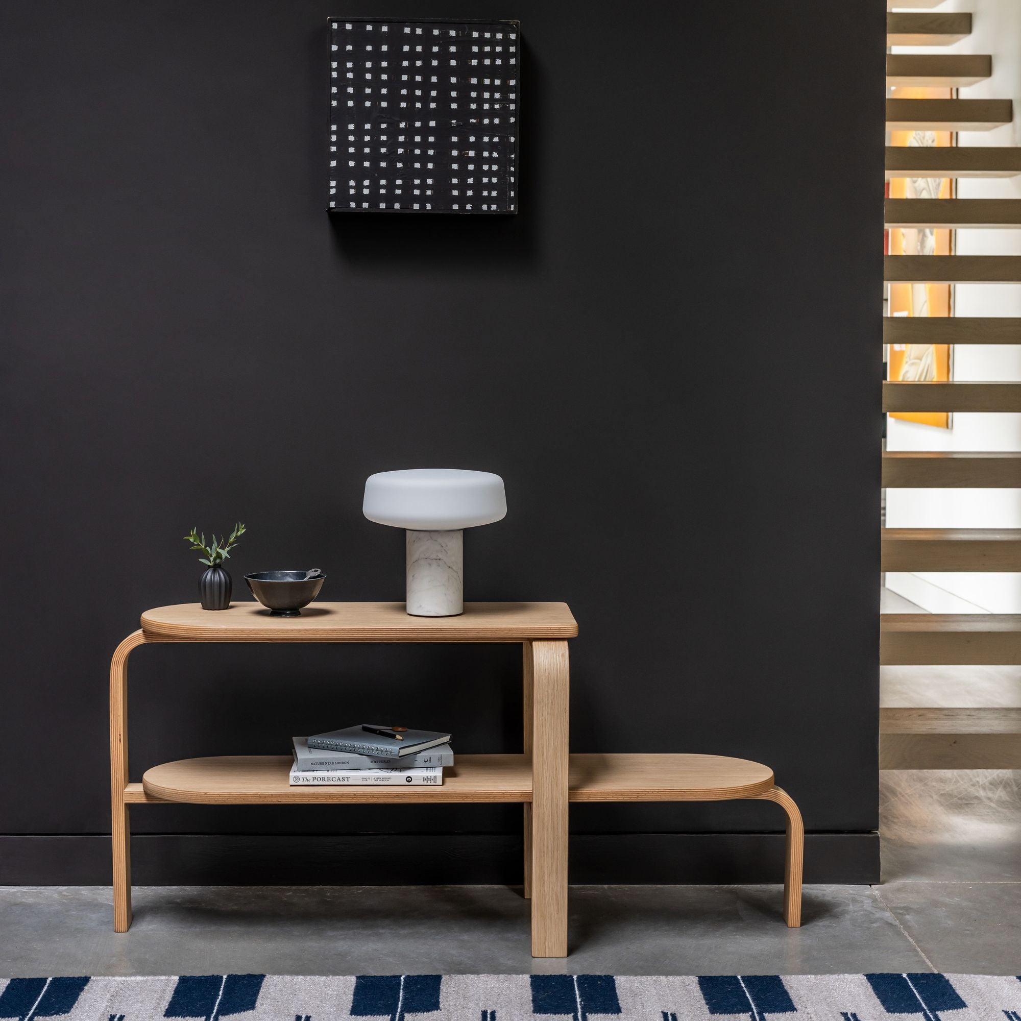 Altura Bench in Oak in a hallway that is painted black with a solid rechargeable light, some books and plant on
